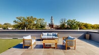 Located at 3624 N Conrad St in Philadelphia's East Falls neighborhood (19129), this remarkable rooftop space offers an expansive outdoor retreat with a thoughtfully designed layout. The rooftop is divided into two distinct functional zones: a generous artificial turf lawn area ideal for recreation and open-air gatherings, and a refined tiled terrace finished in large-format porcelain pavers, perfect for lounging and entertaining. The terrace area is furnished with teak and wicker outdoor seating, providing a polished setting for relaxation under open skies. Perimeter paneled parapet walls in a neutral gray finish define the space while offering privacy and wind protection. A dedicated rooftop access structure with a white door provides convenient entry. The rooftop is surrounded by sweeping panoramic views, highlighted by a striking sightline to the historic East Falls clock tower and surrounding tree canopy. Additional neighborhood rooftops and lush mature trees frame the horizon in every direction, creating a verdant urban backdrop. Abundant natural light floods the space throughout the day, making it a sun-drenched outdoor destination well-suited for both quiet enjoyment and social gatherings in the heart of Philadelphia.