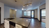 This single-floor residence presents a thoughtfully designed open-concept layout where the kitchen flows seamlessly into the living area. The kitchen serves as a central hub, featuring a large island with dark granite countertops, white cabinetry, a decorative tile backsplash, and a full suite of stainless steel appliances including a French door refrigerator, electric range, built-in microwave, and dishwasher. Louvered closet doors provide additional storage nearby. Track lighting and pendant fixtures illuminate both the kitchen and living spaces, while a ceiling fan enhances comfort in the main living area. Natural light enters through windows fitted with horizontal blinds, and a glass-paneled door provides exterior access. Rich hardwood-style flooring extends throughout the main living spaces, contrasting with the dark trim and door frames that lend a contemporary edge to the interior. The bedroom features neutral carpet and a window for natural light. The bathroom is finished with large-format grey tile flooring, a soaking tub with tiled surround, a dual-sink vanity with dark countertops, white cabinetry, and a wide mirror above. The overall aesthetic is modern and cohesive throughout.