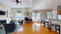 Located at 2350 Freedom Blvd in Florence, South Carolina, this single-story community clubhouse offers a welcoming and well-appointed gathering space designed for both relaxation and productivity. The open-plan layout is anchored by warm hardwood flooring that flows seamlessly throughout, complemented by soft neutral walls and crown molding that lend a polished, cohesive feel.

The main lounge area features soaring tray ceilings with a ceiling fan and recessed lighting, creating an airy atmosphere. Plush teal armchairs with bright yellow accent pillows are arranged around a coffee table near a classic white-mantled fireplace, above which a flat-screen television is mounted. A built-in bookcase and a vanity-style console table provide additional utility and charm.

Large windows along the perimeter invite abundant natural light and offer pleasant views of the surrounding tree-lined grounds. A dedicated workstation with a computer and printer sits conveniently within the lounge, making the space equally suited for focused tasks.

The kitchen area is well-equipped with warm wood cabinetry, granite countertops, stainless steel appliances including a refrigerator and microwave, and a peninsula bar with counter-height seating. Pendant lighting above the bar adds a refined touch. The overall ambiance is bright, functional, and inviting — ideal as a shared community amenity space.