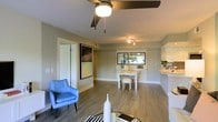 This well-appointed single-floor space offers a thoughtfully designed open-concept layout that connects the living, dining, and kitchen areas in a natural, flowing arrangement. The living area features warm-toned luxury vinyl plank flooring that extends throughout, providing a clean and cohesive foundation for the space. A ceiling fan with an integrated light fixture anchors the main living area, while a track lighting system in the dining zone adds focused, adjustable illumination.

The dining area is defined by a crisp white dining set positioned beneath a decorative wall-mounted mirror that reflects natural light and creates a sense of added depth. A pass-through opening connects the dining space to the kitchen, which features white cabinetry and light countertops, contributing to the bright, airy feel of the overall space.

The neutral gray wall tones provide a versatile backdrop that complements both the warm flooring and the white trim and baseboards, lending the interior a modern yet inviting character. A doorway visible from the main living area hints at additional private rooms beyond. Natural light filters in subtly, enhancing the open, comfortable ambiance. Overall, the space balances functional design with clean, contemporary aesthetics suitable for everyday living.