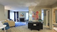 This single-floor space offers a welcoming and thoughtfully designed interior that blends comfort with artistic flair. The main living area is open and spacious, anchored by a classic fireplace with a clean white mantel that serves as a natural focal point. Large windows dressed in rich navy curtains allow generous natural light to filter in, brightening the neutral, carpeted floors throughout.

The space is elevated by bold, colorful artwork displayed prominently on the walls — including a vibrant large-scale multicolored composition that draws the eye and adds a dynamic energy to the room. A dark console table provides functional surface space and serves as a stylish display area for decorative accents, including a sculptural glass bowl and decorative boxes.

A patterned blue area rug defines the seating zone within the living area, complementing the overall color palette and adding visual texture to the space. The warm beige tones of the walls create a neutral backdrop that allows the colorful decor elements to stand out. An adjacent area visible through a doorway suggests additional rooms, hinting at a well-connected and functional floor plan. Overall, the space exudes a lively yet comfortable ambiance, ideal for both relaxing and entertaining.