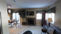 This single-floor residence offers a comfortable and well-appointed living space designed for both relaxation and everyday functionality. The living room serves as the heart of the home, anchored by a prominent stone fireplace with a slate tile hearth that adds a rustic, natural character to the space. A wall-mounted flat-screen television is positioned above the fireplace, creating a practical and visually balanced focal point. Warm, neutral-toned walls complement the carpeted flooring throughout, contributing to a cozy and inviting ambiance. The room benefits from multiple windows dressed with decorative valances, allowing natural light to filter in from several directions. Large indoor plants add a touch of greenery and life to the interior. Adjacent to the living area, a dining space is visible near a sliding glass door, which provides access to an outdoor area and further brightens the room with natural light. A wooden display cabinet with glass doors adds a classic decorative element, offering storage and display space. The overall layout of this single-story home flows naturally between the living and dining areas, making it a functional and comfortable environment suited to a variety of lifestyles.