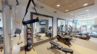 Located at 5659 W Eighth Street in Los Angeles, this dedicated fitness center offers a comprehensive and thoughtfully organized workout environment across a single floor. The space features a neutral color palette with warm beige tones, low-profile recessed and coffered ceiling lighting, and durable wood-look flooring that gives the room a clean, professional feel.

The cardio section is anchored by multiple Life Fitness treadmills positioned beside large windows that frame lush greenery outside, providing natural light and pleasant outdoor views during workouts. Additional cardio equipment includes elliptical trainers and a recumbent cycle. For strength training, the facility offers a well-stocked three-tier dumbbell rack with a full range of free weights, a cable pulley system, and a variety of resistance-based machines. A medicine ball rack adds functional training options to the mix.

Large framed mirrors are mounted along the walls, enhancing the sense of space and allowing users to monitor their form throughout their workout. Multiple wall-mounted flat-screen televisions are positioned throughout the room, ensuring visibility from virtually every station. A glass entry door connects the fitness center to adjacent common areas, contributing to an open and accessible atmosphere. The overall design is clean, functional, and well-maintained, making it a fully equipped facility suited for a wide range of fitness goals.