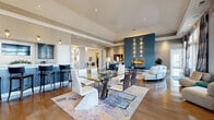 This expansive single-floor amenity facility offers a thoughtfully designed layout across eight distinct areas, delivering a premium community experience. The grand entrance rotunda sets an impressive tone, featuring a soaring coffered ceiling, sculptural pendant lighting in blue and white tones, large arched windows, and a striking central table anchored by a deep-blue patterned area rug.

The open-plan social lounge combines a dining area, living space, and a wet bar/kitchenette into one cohesive environment. The dining zone features a glass-top table with contemporary seating, while a bold teal accent wall with an integrated linear fireplace and wall sconces defines the living area. The kitchenette is appointed with dark cabinetry, a mosaic tile backsplash, a white quartz countertop, and bar-height seating.

A dedicated lounge with hardwood floors, tall windows, and a tray ceiling provides a more intimate retreat, while a casual sitting area with a stone fireplace, wicker furnishings, and blue-gray walls offers a relaxed, residential feel. The fitness center is equipped with cable machines and cardio equipment, benefiting from direct outdoor access and natural light. A bright game room lined with floor-to-ceiling windows houses a foosball table and gaming tables. A tiled transitional corridor with seating and curated artwork connects the spaces seamlessly.