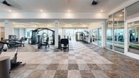 Located in St. Petersburg, Florida, this expansive fitness center is a thoroughly equipped, single-floor facility designed to support a wide range of workout disciplines. The space is defined by its modern architectural aesthetic, featuring crisp white ceilings accented by striking angular linear LED lighting that runs across the ceiling in geometric patterns, adding both style and bright, even illumination throughout.

The main workout floor is a generously proportioned open area finished with a neutral checkered tile pattern, providing ample room for a comprehensive array of strength machines, cable systems, multi-functional training rigs, and free-weight benches. A dedicated functional training zone offers turf flooring, resistance systems, and plyometric equipment, while a separate cycling studio features light wood flooring, bold teal accent walls, full-length mirrors, and a large TV display.

Floor-to-ceiling windows line the perimeter, flooding the space with natural light and offering scenic waterfront and cityscape views of the surrounding Saint Petersburg area. Ceiling fans are positioned throughout to ensure consistent airflow. The cardio zone lines the window wall, positioning treadmills, ellipticals, and stair climbers to take full advantage of the outdoor views. A welcoming entry corridor with glass-panel partitions, elevator access, and curated wall art completes the facility's polished, contemporary feel.