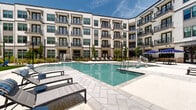 Located at 4850 Rouzan Square Ave in Baton Rouge, Louisiana, this outdoor amenity courtyard is the centerpiece of an upscale residential community, thoughtfully designed to deliver a resort-style living experience. The space is framed on all sides by a striking multi-story building facade featuring white siding, dark-trimmed windows, and private balconies that overlook the courtyard below.

The focal point of the outdoor area is a generously sized swimming pool with crystal-clear blue water, finished with a light stone tile surround that extends into a broad sun deck. Stainless steel entry rails and a shallow wading ledge provide easy pool access, while rows of lounge chairs positioned along the perimeter invite residents to relax in the Louisiana sunshine.

Shaded seating areas equipped with navy-and-white fringed market umbrellas and modern outdoor dining sets offer a comfortable retreat from the sun. Thoughtfully placed landscaping, including ornamental grasses, young trees, and manicured green lawn sections, adds texture and a natural softness to the hardscaped environment. Brick accent columns and wrought-iron fencing define intimate garden pockets near ground-level entries, enhancing the sense of community and privacy. The overall ambiance is sophisticated yet welcoming, blending classic Southern architectural character with contemporary outdoor design.