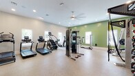 This fitness center offers a spacious, single-floor layout designed to accommodate a full range of workout routines. The space is divided into distinct functional zones, seamlessly connected by durable speckled flooring that extends throughout. Neutral beige walls are complemented by a bold sage green accent wall, adding visual energy to the environment. Recessed lighting and ceiling fans ensure the space remains well-lit and comfortable during use.

The cardio zone is generously equipped with treadmills, a stair climber, an elliptical, a stationary bike with an interactive display, and a rowing machine, all positioned near windows fitted with blinds that allow natural light to filter in. The strength training area features a multi-station cable machine, a power rack, a full dumbbell rack with an assortment of weights, medicine balls, resistance tools, and an adjustable bench. Floor-to-ceiling mirrors line one wall, enhancing the sense of space and aiding in form correction.

A wall-mounted flat-screen television is positioned for visibility across the main floor. The entry area includes a sanitizing station and posted fitness center guidelines, reflecting a clean, well-maintained environment.