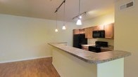 This single-floor residence is designed with an open, functional layout that promotes a seamless flow between the kitchen and living areas. The kitchen serves as the heart of the space, anchored by a generous wraparound breakfast bar with a light stone-toned countertop that doubles as both a food preparation surface and a casual dining area. Warm medium-toned wood cabinetry lines the kitchen walls, complemented by a decorative wine rack detail that adds a touch of character. Black appliances, including a refrigerator, microwave, range, and oven, provide a sleek, cohesive aesthetic throughout the kitchen. Overhead, a combination of track lighting and elegant pendant lights illuminates the space with a warm glow, highlighting both the countertops and the open layout beyond. The flooring throughout is a rich hardwood-style surface that adds warmth and continuity from the kitchen into the adjoining living area. Neutral, light-toned walls keep the space feeling bright and open, providing a versatile backdrop for a range of interior styles. The single-level layout ensures easy navigation throughout, making the space both practical and inviting for everyday living.