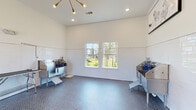 This single-floor pet grooming facility is thoughtfully designed to provide a functional and comfortable environment for both pets and their caretakers. The room features white-painted walls accented by a wainscoting-height band of glossy white subway tile, creating a clean, sanitary aesthetic throughout the space.

The flooring consists of blue-gray penny tile, which adds texture and visual interest while providing a durable, easy-to-clean surface. A floor drain is centrally located, underscoring the room's practical design. Ceilings rise generously overhead and are illuminated by a combination of recessed lighting and a striking sputnik-style chandelier with gold accents, lending a modern touch to the utilitarian setting.

Multiple stainless steel wash tubs are positioned along the walls, each equipped with steps for pet access and grooming supplies. Adjustable grooming tables with overhead arm attachments allow for versatile use, while a wall-mounted pet dryer adds further convenience. Natural light enters through several double-hung windows offering outdoor views of a green landscaped area. Framed pet-themed artwork decorates the upper walls, and a clearly marked fire extinguisher and posted rules signage reflect attention to safety and organization. A dedicated entry door provides direct exterior access.