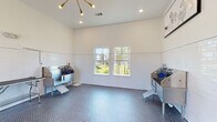 This single-floor pet grooming facility is thoughtfully designed to provide a functional and comfortable environment for both pets and their caretakers. The room features white-painted walls accented by a wainscoting-height band of glossy white subway tile, creating a clean, sanitary aesthetic throughout the space.

The flooring consists of blue-gray penny tile, which adds texture and visual interest while providing a durable, easy-to-clean surface. A floor drain is centrally located, underscoring the room's practical design. Ceilings rise generously overhead and are illuminated by a combination of recessed lighting and a striking sputnik-style chandelier with gold accents, lending a modern touch to the utilitarian setting.

Multiple stainless steel wash tubs are positioned along the walls, each equipped with steps for pet access and grooming supplies. Adjustable grooming tables with overhead arm attachments allow for versatile use, while a wall-mounted pet dryer adds further convenience. Natural light enters through several double-hung windows offering outdoor views of a green landscaped area. Framed pet-themed artwork decorates the upper walls, and a clearly marked fire extinguisher and posted rules signage reflect attention to safety and organization. A dedicated entry door provides direct exterior access.