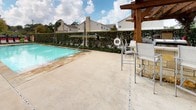 This outdoor amenity space centers around a large, sparkling swimming pool with a generously sized concrete deck designed for relaxation and recreation. A row of lounge chairs with red accent cushions lines the poolside, offering plentiful space for sunbathing along the full length of the pool. Vibrant red umbrellas are strategically positioned throughout the deck, providing shaded seating areas with tables and chairs for casual gatherings. A handsome wooden pergola anchors one end of the pool area, adding architectural character and additional shade. The space is enclosed by a decorative iron fence and features a gated entry accented by a warm wood privacy wall and lush potted greenery, including tropical palms and leafy plants that enhance the resort-like atmosphere. A safety ring is mounted poolside, and the pool itself is equipped with water features for added enjoyment. The surrounding landscape includes mature trees that frame the space and offer a natural backdrop. The open-air design allows for abundant natural light and open sky views, creating a bright and inviting environment. The overall ambiance is that of a community retreat, thoughtfully designed for outdoor leisure and social activity.