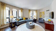 This single-floor residence is thoughtfully designed with a contemporary aesthetic, showcasing a consistent color palette of soft grey, warm yellow, and neutral tones throughout. A welcoming entry hallway with wood-look flooring and recessed lighting leads into the main living areas.

The living room is a standout space, featuring multiple large windows dressed with patterned yellow curtains and roller shades that invite generous natural light and urban views. A curved sectional sofa, plush white area rug, and cylindrical ottoman anchor the room, while wall-mounted artwork adds a decorative focal point. The space flows naturally to a wall-mounted television area with additional seating.

The kitchen is well-appointed with rich cherry wood cabinetry, a dark subway tile backsplash, light countertops, and a full suite of stainless steel appliances including a refrigerator, range, microwave, and dishwasher. A freestanding island provides added prep and storage space. Wood-look flooring ties the kitchen to the main living area seamlessly.

Two bedrooms offer carpeted floors, oversized windows with urban views, and ample closet space, including a walk-in closet in one room. The bathroom features tile flooring, a wood vanity with light countertops, and clean, neutral finishes.