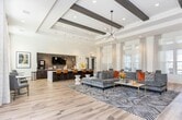 Located at 2021 Bluff Creek St in El Paso, Texas, this single-story community clubhouse offers a thoughtfully designed gathering space that blends contemporary style with functional amenities. The open-concept layout is anchored by soaring ceilings accented with dark exposed beams, recessed lighting, and light wood-look flooring that flows seamlessly throughout the entire space. Floor-to-ceiling windows dressed with sheer white drapery flood the interior with abundant natural light, creating a bright and welcoming atmosphere. The lounge area features a generously sized sectional sofa in a rich gray upholstery, complemented by bold accent pillows and a striking geometric-patterned area rug. A prominent bar and counter space serves as a central hub, finished with a luxurious white marble-style countertop, rich dark cabinetry, and a row of cognac leather barstools. Pendant lighting with teardrop glass fixtures hangs above the counter, adding an elegant finishing touch. A wall-mounted flat-screen TV and a Starbucks-branded coffee station enhance the space's social and functional appeal. Additional bistro-style seating near the windows offers quieter spots for conversation or individual use. The entrance foyer features a warm terracotta accent wall and decorative planters, setting an inviting tone from the moment of arrival.