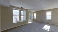 This single-floor space offers a clean and functional layout designed to maximize comfort and natural light. The main living area is a standout feature, characterized by its generous proportions and an abundance of natural light streaming through a large multi-pane window and an additional side window. These windows frame views of the surrounding urban environment, including neighboring brick buildings and a city skyline in the background, providing a dynamic connection to the outside world.

The interior presents a neutral color palette, with warm beige walls and white trim that create a calm and versatile backdrop suited to a variety of furnishing styles. Soft, plush carpeting in a muted gray tone covers the floors throughout, adding warmth and comfort underfoot. The ceiling is smooth and finished in white, with a centrally located HVAC vent ensuring year-round climate comfort.

The overall ambiance of the space is understated and practical, with clean lines and minimal architectural detailing that allow for flexible use and personalization. The open feel of the main living area, combined with ample natural light and urban views, makes this a welcoming and livable space well-suited for everyday life in an active urban setting.