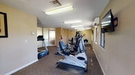 Located at 6942 E Rosewood Cir in Tucson, Arizona, this single-floor fitness center is designed to provide a functional and comfortable workout environment. The space is finished with warm beige walls, textured ceilings, and durable carpet flooring throughout, creating a practical yet welcoming atmosphere.

The facility is well-equipped with a variety of cardio machines, including multiple elliptical trainers, a recumbent bike, and a Spirit treadmill. A multi-station strength training machine is positioned toward the rear of the room, offering a full range of resistance-based exercises. A wall-mounted flat-screen television provides entertainment during workouts.

Natural light enters through multiple windows along the exterior walls, offering views of the surrounding Tucson landscape. A wall-mounted mini-split air conditioning unit ensures a comfortable temperature year-round. The space is also equipped with a fire extinguisher and security cameras, reflecting attention to safety.

Frosted glass panel partitions near the entrance add a subtle architectural detail while providing a degree of privacy. Decorative framed artwork and a wall clock contribute to the overall tidiness of the space. Fluorescent overhead lighting ensures the entire facility is brightly and evenly illuminated, supporting a safe and effective workout experience at any time of day.