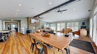 Located at 5 Fall Wood Dr in Hamilton, Ohio, this single-floor community gathering space offers a thoughtfully designed open-concept layout that seamlessly connects multiple functional areas. The living room serves as the centerpiece, anchored by a striking floor-to-ceiling stacked stone fireplace topped with a mounted display. Exposed dark ceiling beams and dual modern ceiling fans add architectural character, while recessed lighting and large French doors with flanking windows flood the space with abundant natural light and views of the surrounding greenery.

Rich hardwood flooring flows throughout, unifying the dining and living zones. The main dining area features a substantial live-edge wood communal table, comfortably seating multiple guests. A secondary dining zone with round marble-top tables and industrial-style chairs provides additional flexible seating. A kitchen bar with a light stone countertop and bar stools connects to a dedicated refreshment station in the background.

A separate room off the main area is outfitted with individual computer workstations, leather chairs, and a round conference table, making it well-suited for focused work or small meetings. Sage green walls, crown molding, and tasteful décor including woven wall accents and botanical greenery contribute to a warm, contemporary ambiance throughout.