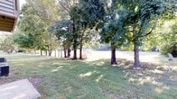 This single-level property is distinguished by its expansive outdoor grounds, which form a central feature of the space. The lot is characterized by a well-maintained lawn that stretches broadly, offering generous open space across the property. A notable row of large, mature trees lines a portion of the grounds, their full canopies providing natural shade and creating a sense of established, park-like character. The interplay of sunlight filtering through the tree cover casts dappled light across the grass, contributing to a calm and natural ambiance throughout the outdoor area. The open lawn beyond the tree line is clearly visible, suggesting a versatile, multi-use outdoor environment suitable for a variety of activities. A structure is partially visible in the background, partially framed by the tree line, indicating the presence of a building on or adjacent to the property. The overall setting conveys a spacious, serene, and well-established outdoor environment with a strong connection to nature, benefiting from abundant natural light and mature landscaping that adds both visual interest and practical shade to the grounds.
