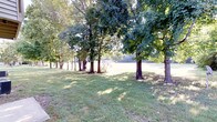 This single-level property is distinguished by its expansive outdoor grounds, which form a central feature of the space. The lot is characterized by a well-maintained lawn that stretches broadly, offering generous open space across the property. A notable row of large, mature trees lines a portion of the grounds, their full canopies providing natural shade and creating a sense of established, park-like character. The interplay of sunlight filtering through the tree cover casts dappled light across the grass, contributing to a calm and natural ambiance throughout the outdoor area. The open lawn beyond the tree line is clearly visible, suggesting a versatile, multi-use outdoor environment suitable for a variety of activities. A structure is partially visible in the background, partially framed by the tree line, indicating the presence of a building on or adjacent to the property. The overall setting conveys a spacious, serene, and well-established outdoor environment with a strong connection to nature, benefiting from abundant natural light and mature landscaping that adds both visual interest and practical shade to the grounds.