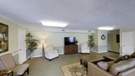 The Media Room is a thoughtfully designed communal space built for comfort and entertainment. The room features soft sage-green walls accented by a white chair rail, creating a calm and inviting atmosphere. Overhead, flush-mounted fluorescent light fixtures provide bright, even illumination across the carpeted floor, which is anchored by a decorative area rug in muted earth tones. At the focal point of the room sits a large flat-screen television atop a rich, dark-wood media console, flanked by a pair of classic upholstered armchairs with elegant carved legs — ideal for casual viewing. A spacious leather sectional sofa is positioned nearby, offering ample seating for groups. Tall potted bamboo plants are placed on either side of the room, adding a touch of natural greenery and softening the overall aesthetic. A decorative wall clock and a traditional table lamp contribute to the warm, homey feel of the space. The textured ceiling adds a classic finishing touch, while the open layout allows for easy movement and social interaction, making this media room equally suited for individual relaxation or group gatherings.