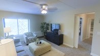 This single-floor space features a living room designed for comfort and practicality. The room is finished with wall-to-wall carpeting in a neutral tone, contributing to a warm and inviting atmosphere. A ceiling fan with an integrated light fixture is centrally mounted, providing both airflow and ambient lighting. Natural light enters through a window fitted with blinds, brightening the space and offering a view of lush greenery outside. The walls are painted in a soft, muted green tone, lending the room a calm and relaxed character. A dark, horizontally ribbed media console serves as a focal point, supporting a flat-screen television and decorative accents. Seating includes a patterned armchair in blue and grey tones, complemented by a bold yellow and white geometric-patterned ottoman that doubles as a coffee table. A tall indoor plant adds a natural, organic element to the corner of the room. White baseboards and trim provide clean contrast against the walls, while a partially visible doorway with a slatted blind hints at additional adjoining space. Overall, the living area presents a tidy, well-appointed environment with a blend of neutral and accent tones.