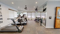 This fitness center offers a comprehensive workout environment within a single, open-plan space. The room is finished with durable patterned carpet tile flooring and features white walls accented by a bold motivational wall mural reading "The Best Project You Will Ever Work On Is You," adding an energizing atmosphere to the space.

Natural light fills the room through multiple windows fitted with horizontal blinds, providing views to the outdoors. Recessed lighting and ceiling fans with light fixtures ensure consistent illumination and air circulation throughout. Large floor-to-ceiling mirrors on one wall visually expand the space and assist users in monitoring their form during exercise.

The facility is well-stocked with a variety of equipment, including multiple treadmills, elliptical machines, and a multi-station cable and weight machine. A dedicated free weights area features a dumbbell rack, weight plates, and a stability ball. A tiered seating bench near the entry provides a space to sit and prepare for workouts.

Near the entrance, a water dispenser, cork bulletin board, and coat hooks serve as practical amenities for users. A warm wood-toned Dutch door marks the entry point. The overall design is functional and motivating, making it a practical and inviting space for a full range of fitness activities.