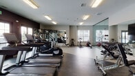 Located at 14750 S Wallin Dr in Plainfield, IL, this well-appointed fitness center is designed to support a comprehensive workout experience within a single, thoughtfully organized floor. The space is divided into distinct functional zones, each tailored to a specific type of training. The main cardio area features a row of Life Fitness treadmills and elliptical machines aligned along a dark-accented accent wall, complemented by large windows that introduce natural light and views of the surrounding exterior. A dedicated strength training section houses a multi-station Life Fitness cable and weight machine system alongside a dumbbell rack, all set against full-length mirrors that visually expand the space. A separate spin and functional training zone includes cycling bikes, a rowing machine, and various free weights and medicine balls. A glass-partitioned studio area with a light-toned floor provides an open, flexible environment for stretching, yoga, or group exercise, equipped with stability balls and resistance equipment. Recessed and overhead fluorescent lighting keeps the entire facility bright and evenly illuminated throughout. The flooring transitions between rubberized gym surfaces and smooth studio flooring, each suited to its intended activity. Glass entry doors connect the facility to adjacent amenity spaces, reinforcing an open and accessible design throughout.