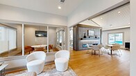 This thoughtfully designed single-floor space offers a generous, open-concept layout that seamlessly integrates multiple functional areas under one roof. The main gathering area is anchored by a striking floor-to-ceiling stone fireplace wall with a linear gas fireplace, a built-in display shelf, and a wall-mounted television — serving as a dramatic focal point for the lounge seating arranged around it. Vaulted ceilings with exposed beams and white shiplap detailing lend architectural character and a sense of spacious airiness throughout, complemented by large ceiling fans and recessed lighting.

The open dining area accommodates multiple seating arrangements, with light wood flooring flowing continuously throughout. Large windows and glass doors offer views of a pool area, flooding the interior with natural light. A well-appointed kitchenette features sage-green cabinetry, a herringbone tile backsplash, stainless countertops, a beverage cooler, and pendant lighting — ideal for hosting or everyday convenience.

Glass-walled private meeting rooms provide semi-enclosed spaces for focused work or small gatherings, each equipped with a wall-mounted screen, pendant lighting, and ample natural light. A separate lounge area with large exterior-facing windows invites relaxed seating with garden views. The overall ambiance is clean, contemporary, and welcoming.