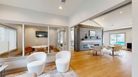 This thoughtfully designed single-floor space offers a generous, open-concept layout that seamlessly integrates multiple functional areas under one roof. The main gathering area is anchored by a striking floor-to-ceiling stone fireplace wall with a linear gas fireplace, a built-in display shelf, and a wall-mounted television — serving as a dramatic focal point for the lounge seating arranged around it. Vaulted ceilings with exposed beams and white shiplap detailing lend architectural character and a sense of spacious airiness throughout, complemented by large ceiling fans and recessed lighting.

The open dining area accommodates multiple seating arrangements, with light wood flooring flowing continuously throughout. Large windows and glass doors offer views of a pool area, flooding the interior with natural light. A well-appointed kitchenette features sage-green cabinetry, a herringbone tile backsplash, stainless countertops, a beverage cooler, and pendant lighting — ideal for hosting or everyday convenience.

Glass-walled private meeting rooms provide semi-enclosed spaces for focused work or small gatherings, each equipped with a wall-mounted screen, pendant lighting, and ample natural light. A separate lounge area with large exterior-facing windows invites relaxed seating with garden views. The overall ambiance is clean, contemporary, and welcoming.