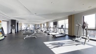 This contemporary fitness center offers a spacious, thoughtfully designed environment for a comprehensive workout experience. The facility is anchored by a generous open floor plan finished with durable dark rubberized flooring, providing both safety and functionality throughout the space.

A full row of treadmills and cardio machines is positioned along the perimeter, taking full advantage of the expansive floor-to-ceiling windows that line one side of the room. These large windows flood the space with abundant natural light and offer sweeping panoramic views of the surrounding cityscape, creating an energizing atmosphere for users.

The strength training area is equally well-appointed, featuring a variety of free weights, benches, and resistance machines arranged to maximize usable floor space while maintaining clear sightlines across the room. A mounted flat-screen television provides entertainment during workouts.

The ceiling is finished with clean, angled panels and recessed lighting that complement the overall modern aesthetic. Warm wood-toned accents along the window columns contrast elegantly with the neutral grey and white palette of the walls and ceiling. The overall ambiance is sleek, bright, and professional, making this fitness center a standout amenity designed to meet the needs of a diverse range of users.