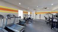 This single-level fitness center offers a dedicated space for cardio and strength training in a bright, energizing environment. The room is finished with light gray walls accented by bold horizontal stripes in orange and yellow, creating a dynamic and motivating atmosphere. Recessed lighting and dual ceiling fans ensure the space remains well-lit and comfortable during workouts.

The flooring consists of durable rubber gym matting, providing cushioning and traction suited for high-traffic exercise use. The cardio section is well-appointed with multiple treadmills, elliptical trainers, and a stationary bike, all positioned to take advantage of the natural light streaming in through the windows along one wall.

Along the perimeter, a medicine ball rack holds a variety of weighted balls for functional training, and additional strength equipment is visible toward the far end of the room. A wall-mounted display adds a finishing touch to the space. The open layout allows for comfortable movement between equipment, and the combination of natural and artificial lighting keeps the environment inviting and functional throughout the day.