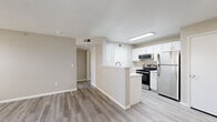 This thoughtfully laid-out single-story unit offers a practical and comfortable living experience across its efficiently organized floor plan. The combined kitchen and living area forms the heart of the home, where light-toned, wood-look luxury vinyl flooring flows seamlessly throughout. The kitchen is outfitted with crisp white cabinetry, stainless steel appliances including a refrigerator, range with microwave above, and a single-basin sink set into clean white countertops. A peninsula-style divider separates the kitchen from the open living area, enhancing the sense of connected space. The living room benefits from a full-light entry door and a wide window fitted with horizontal blinds, filling the room with natural light and offering views of greenery outside. A hallway connects the main living areas to the private quarters, where the bedroom features plush carpeting, a ceiling fan with integrated lighting, and a window that continues the theme of natural illumination. A dedicated closet provides practical storage. The bathroom is clean and functional, complete with a full tub-shower combination, toilet, and vanity with wood-look flooring. The entry hallway offers additional transition space, rounding out this well-appointed, move-in ready unit.