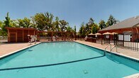 Located on Park Lane in Dunellen, New Jersey, this outdoor amenity space is designed to serve as a central gathering and recreation hub. At its heart is a generously sized swimming pool with clear, turquoise water, framed by a well-maintained tiled deck. The pool area is equipped with designated depth markers and safety features, reflecting a thoughtfully designed aquatic environment.

Surrounding the pool, the expansive patio features ample lounge seating, including rows of reclining chairs and umbrella-shaded tables, providing both sun and shade options for residents and guests. Multiple pergola structures add architectural interest while creating comfortable, shaded gathering spots throughout the deck.

A single-story brick clubhouse anchors one side of the amenity area, characterized by its classic red brick facade, gabled roofline, large windows, and a welcoming central entrance. The building opens directly onto the stone-paved patio, seamlessly connecting indoor and outdoor spaces.

The entire pool and deck area is enclosed by a decorative metal fence, ensuring security while maintaining open sightlines to the surrounding residential buildings and mature tree canopy. Under clear blue skies, the space is bathed in natural light, creating a bright, resort-like atmosphere ideal for outdoor recreation and relaxation.