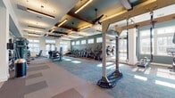 This fitness center offers a comprehensive workout environment designed to support a wide range of training needs. The space is anchored by a row of cardio machines — including treadmills, ellipticals, and stair climbers — positioned along a teal-accented accent wall beneath a bank of transom windows that flood the floor with natural light. Ceiling fans and linear pendant lighting are integrated into a coffered ceiling with dark wood beam details, lending warmth and architectural character to the space. A dedicated functional training area features a multi-station rig outfitted with battle ropes, TRX suspension straps, kettlebells, medicine balls, and a heavy punching bag, ideal for high-intensity and cross-training workouts. Strength training stations and cable machines with mirrors line one side of the facility, providing ample options for resistance work. Patterned carpet tiles in muted blue and gray tones define different zones throughout the floor plan. Large glass double doors open to outdoor views, enhancing the sense of openness and connectivity to the surrounding community. A water bottle filling station set against a textured tile wall provides a practical, polished amenity. Overall, the fitness center balances modern design with functional versatility.
