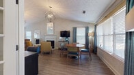 This single-floor space combines an open, multi-purpose layout with warm, contemporary design elements. The main living and dining area is anchored by vaulted ceilings with a textured finish, drawing the eye upward to a striking geometric chandelier that serves as the room's central focal point. Wide-plank wood-look flooring runs throughout, unifying the space with a clean, cohesive aesthetic.

The living area features a fireplace with a classic white surround, a wall-mounted flat-screen television atop a media console, and a comfortable seating arrangement with accent chairs in bold mustard and teal tones. Adjacent to the living space is a round dining table with upholstered chairs, positioned near a bank of large windows dressed in teal drape panels and white blinds that allow an abundance of natural light to fill the room.

A dedicated workstation lines one wall, equipped with a long desk surface, computer monitor, and rolling chairs — ideal for productivity. Beyond this, a kitchen area features a raised bar with cushioned stools and full appliances, including a refrigerator and ample counter space.

Near the entry, a reception-style console with a round mirror and flanking lamps serves as a welcoming focal point. Throughout the space, curated artwork and layered lighting contribute to a warm, inviting, and well-appointed atmosphere.