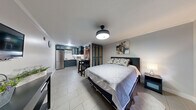Located at 2211 Paradise Rd in Las Vegas, NV, this efficiently designed studio unit combines bedroom, living, dining, and kitchen functions within a single open-plan layout. Light-toned large-format tile flooring runs throughout, creating a cohesive and airy feel. The sleeping area accommodates a full-sized bed complemented by a dark wood headboard, coordinating nightstand, and wall-mounted artwork, while a barn-style sliding door leads to a dedicated closet with teal-painted paneled storage. Warm natural wood accents frame a front-facing window with a roller shade and a solid wood entry door, adding character to the space. A wall-mounted flat-screen TV and dresser complete the living side of the room. The kitchen stands out with bold teal shaker-style cabinetry, white marble-look countertops, a classic subway tile backsplash, stainless steel appliances including a full-sized refrigerator, and a black matte faucet. A compact bar-height counter with seating provides a casual dining option. The bathroom features a striking glass-enclosed walk-in shower clad in rich, earth-toned stone-look tile, a glass vessel sink atop a marble vanity, and a small operable window for ventilation. A modern flush-mount ceiling light anchors the main room, enhancing the contemporary ambiance of this thoughtfully appointed Las Vegas unit.