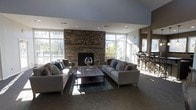 This single-floor space is designed with an open, airy layout that immediately draws attention to its most prominent feature: a floor-to-ceiling stacked stone fireplace serving as the focal point of the main lounge area. The fireplace wall is flanked by large, multi-pane windows that flood the space with generous natural light and offer views of the surrounding outdoor environment, creating a bright and welcoming atmosphere throughout the day.

The lounge area is furnished with two neutral-toned sofas accented with patterned throw pillows in blues and earth tones, arranged around a dark wood coffee table atop a decorative area rug. A large decorative clock is mounted above the fireplace mantle, adding a stylish finishing touch.

The open floor plan connects the lounge seamlessly to an adjacent area visible to the right, which features bar-height seating and darker furnishings, suggesting a dining or bar space. The combination of stone, light walls, and wood tones gives the overall space a contemporary yet warm aesthetic suited for gathering and relaxation.