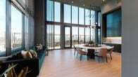 This elevated sky lounge is a thoughtfully designed amenity space that blends modern architecture with upscale finishes across a single floor. The centerpiece is a dramatic double-height great room, where floor-to-ceiling windows and glass doors frame sweeping panoramic city views, flooding the interior with natural light. Sheer textured drapes frame the towering glazing, while light wood-toned flooring and dark charcoal walls create a refined contrast. A sculptural globe chandelier anchors the dining area, which features a round wood table surrounded by mixed seating. A deep emerald green sectional sofa provides a comfortable lounge corner adjacent to the windows and balcony access.

The kitchenette is outfitted with teal cabinetry, brass hardware, white stone countertops, a marble-style backsplash, and integrated Bosch appliances. A quieter seating alcove with carpeted floors and floor-to-ceiling windows offers an intimate retreat with cityscape views. A media lounge includes a marble-topped bar-height counter, wall-mounted TV, and curated artwork. The space also features a Full Swing golf simulator room with a dedicated hitting mat, projection screen, and equipment storage, rounding out this versatile and amenity-rich environment.