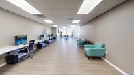 Located at 919 N Stone Ave in Tucson, Arizona, this single-floor commercial space offers a clean, modern environment designed to support a variety of work and collaborative activities. The layout is open and elongated, finished with light wood-style flooring and neutral warm-gray walls that create a bright, welcoming atmosphere throughout.

A dedicated computer workstation area runs along one wall, featuring a long continuous desk with multiple desktop computers and ergonomic high-back office chairs — well-suited for productivity or shared computing needs. Colorful abstract artwork accents the wall above the workstations, adding a touch of creative energy to the space.

Adjacent to the workstations, a comfortable lounge area is furnished with vibrant teal upholstered chairs and sleek side tables, providing an inviting spot for casual meetings or relaxed work sessions. Toward the far end of the space, a wall of large windows floods the area with abundant natural light and offers views of the surrounding Tucson streetscape and greenery. This windowed zone includes high-top tables and bar-height stools, ideal for collaborative gatherings or informal discussions.

Recessed panel lighting in a drop ceiling provides consistent, even illumination throughout. The space is further accented with potted greenery near the windows, enhancing the fresh, modern feel. A private door at the rear provides additional access or utility. Overall, the space delivers a functional yet comfortable professional environment.