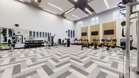 Located at 900 Crest Ln in Fort Lee, NJ, this fitness center is a thoughtfully designed, single-floor facility that blends modern aesthetics with comprehensive workout functionality. The space is defined by its striking double-height ceilings, which lend an open, airy atmosphere and are complemented by large clerestory windows that draw in abundant natural light from multiple angles.

The flooring throughout features a distinctive herringbone-patterned carpet tile in varying shades of gray and white, adding a refined, contemporary feel to the space. Suspended rectangular lighting panels provide even, bright illumination across the entire floor.

The facility is divided into functional workout zones. A dedicated cardio area lines one wall, outfitted with treadmills, stationary bikes, rowing machines, and ellipticals—each equipped with individual display screens. Flat-screen televisions mounted on warm wood slat accent panels enhance the cardio experience. A strength training zone includes cable machines, a free weight rack with dumbbells, adjustable benches, and a power rack. A separate turf area supports functional training with medicine balls and resistance equipment. Decorative acoustic panels in a mosaic of blue and gray tones adorn the walls, adding both visual interest and sound management to this well-appointed fitness facility.