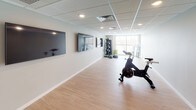 This single-floor fitness studio is thoughtfully designed to support a range of workout activities in a clean, modern environment. The space features light wood laminate flooring throughout, providing a durable yet attractive surface well-suited to physical activity. The drop-tile ceiling incorporates recessed lighting and integrated HVAC vents, ensuring consistent illumination and climate control across the room.

The main workout area is generously proportioned, offering ample open floor space for group fitness classes or individual training sessions. Multiple large flat-screen televisions are mounted along one wall, creating an engaging atmosphere for instructor-led or media-driven workouts. Equipment on hand includes spin bikes, kettlebells, step platforms, medicine balls, resistance tools, and tiered dumbbell racks with a wide weight range.

A large window at one end of the studio allows natural light to fill the space and offers views of the surrounding outdoor landscape. Entry is provided through a sleek dark-framed glass door with a sidelight panel, adding a polished, professional feel to the studio. Neutral gray walls and white baseboards contribute to a clean, distraction-free aesthetic throughout.