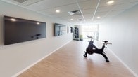 This single-floor fitness studio is thoughtfully designed to support a range of workout activities in a clean, modern environment. The space features light wood laminate flooring throughout, providing a durable yet attractive surface well-suited to physical activity. The drop-tile ceiling incorporates recessed lighting and integrated HVAC vents, ensuring consistent illumination and climate control across the room.

The main workout area is generously proportioned, offering ample open floor space for group fitness classes or individual training sessions. Multiple large flat-screen televisions are mounted along one wall, creating an engaging atmosphere for instructor-led or media-driven workouts. Equipment on hand includes spin bikes, kettlebells, step platforms, medicine balls, resistance tools, and tiered dumbbell racks with a wide weight range.

A large window at one end of the studio allows natural light to fill the space and offers views of the surrounding outdoor landscape. Entry is provided through a sleek dark-framed glass door with a sidelight panel, adding a polished, professional feel to the studio. Neutral gray walls and white baseboards contribute to a clean, distraction-free aesthetic throughout.