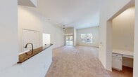 This single-story residence offers a bright, open-concept design characterized by soaring vaulted ceilings and a neutral color palette that enhances the sense of airiness throughout. The spacious living area features plush wall-to-wall carpeting and is anchored by a ceiling fan overhead. Large sliding glass doors and windows flood the space with natural light while providing views of the surrounding greenery. The layout flows seamlessly into the kitchen, which is equipped with white cabinetry, dark granite-style countertops, a peninsula with a built-in sink, hardwood-style flooring, and white appliances including a gas range, microwave, dishwasher, and refrigerator. Recessed lighting adds a polished finish to the kitchen area. The bedrooms are carpeted and well-lit by windows with outdoor views. One bedroom benefits from multiple windows on adjoining walls, maximizing natural light. A generously sized walk-in closet serves the primary bedroom, outfitted with double hanging rods and built-in shelving. The bathrooms include a vanity with a large mirror and ample cabinet storage, as well as a tiled soaking tub and shower combination, completing this well-appointed residence.
