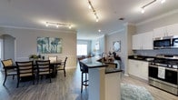 This single-floor residence offers a thoughtfully designed open-concept layout that seamlessly connects the kitchen, dining, and living areas into one cohesive and functional living space. The kitchen is outfitted with crisp white cabinetry, dark granite countertops, and a central island with a built-in sink and bar seating — ideal for casual dining or entertaining. Track lighting runs along the ceiling, providing focused, adjustable illumination that complements the recessed lighting throughout. Crown molding adds a refined architectural detail that frames each room with a classic touch. The dining area accommodates a full-sized table and chairs, anchored by a vibrant piece of wall art that adds a pop of color to the neutral palette. Light gray walls and wood-look flooring run throughout the space, creating a warm yet contemporary ambiance. A glimpse into the adjoining living area reveals a naturally lit room with additional seating, offering a comfortable retreat. The overall design balances practicality with aesthetic appeal, making it a welcoming and versatile space suited for both everyday living and hosting guests.