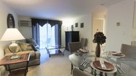 This single-floor residence showcases an open-plan design that integrates the dining and living areas into one cohesive, flowing space. The dining area is anchored by a round glass-top table surrounded by upholstered chairs in a neutral tone, elegantly set with dinnerware and accented by a decorative floral arrangement in a dark vase — adding a sophisticated touch to the space.

The living area extends naturally from the dining zone, featuring a comfortable sofa with decorative pillows and a sleek glass coffee table resting on plush carpeting. A dark armoire serves as a functional focal point along one wall, providing both storage and visual contrast against the light-toned walls and ceiling.

A large sliding glass door dressed with dramatic dark curtains and valance draws the eye toward an outdoor balcony, flooding the interior with natural light and offering a sense of openness. The balcony, visible through the glass, provides additional outdoor living potential.

Throughout the space, the neutral color palette is complemented by carefully chosen dark accent pieces, creating a balanced and inviting ambiance suited for both everyday living and entertaining.