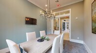 This elegantly appointed space is organized into two distinct functional areas, each thoughtfully designed with high-end finishes and cohesive décor. The conference and dining room serves as the centerpiece, featuring a substantial light wood table surrounded by white upholstered chairs, all anchored by a sophisticated geometric chandelier with candle-style lights. Soft sage-green accent walls complement the neutral crown molding and white trim, while a wall-mounted flat-screen television and large-scale abstract artwork add purposeful visual interest. A floor-to-ceiling window floods the room with natural light and frames views of lush greenery beyond, enhancing the room's welcoming ambiance. A plush area rug grounds the seating arrangement, and French doors with transom windows connect this room to the adjacent space. The business center area features large-format light tile flooring, a continuous granite-topped workstation with built-in computer workstations, and ample task seating. This area is adorned with a pair of striking oversized oyster paintings, lending an artistic coastal sensibility to the professional environment. Decorative metal wall sculptures and fresh floral arrangements add warmth, while glass-panel entry doors allow natural light to flow through the corridor, creating a bright and inviting atmosphere throughout.