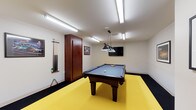 This single-floor game room is thoughtfully designed as a dedicated recreational space, offering an engaging environment for leisure and entertainment. At the heart of the room sits a full-size pool table with a blue felt surface and a dark wood frame, positioned beneath a stylish multi-shade pendant light that provides focused illumination for gameplay. The bold yellow floor area beneath the table creates a striking visual contrast against the darker border flooring, giving the space a lively, dynamic character. A wall-mounted cue rack keeps several pool cues neatly organized and within easy reach, while a wooden storage cabinet offers additional functionality. Framed artwork and posters adorn the white walls, adding personality and a sense of casual charm. A flat-screen television is mounted on the wall opposite the table, making the room suitable for watching sports or entertainment between games. Overhead fluorescent lighting ensures the entire space is well-lit, complementing the pendant fixture above the table. The overall ambiance is energetic yet functional, striking a balance between a casual rec room and a well-equipped entertainment space ideal for socializing and friendly competition.