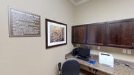 Located at 4500 Highway 57 in Ocean Springs, Mississippi, this business center offers a well-organized and functional environment designed to meet a variety of professional and productivity needs. The single-floor space is finished with warm beige walls, crown molding, and neutral carpeting throughout, creating a polished yet comfortable atmosphere.

The main workstation area features an L-shaped granite countertop workspace lined with dark espresso-stained upper cabinetry for ample storage. Multiple computer terminals, keyboards, and printers — including both laser and multifunction units — are readily available, making this a fully equipped hub for printing, scanning, and computing tasks. Ergonomic rolling chairs provide seating at each station, and posted business center rules ensure a respectful shared environment.

A secondary windowed room offers additional workspace with built-in granite-topped desks along opposing walls and upper cabinetry above. Natural light filters in through a window dressed with horizontal blinds and a striped valance, and framed nature-inspired artwork adds a decorative touch throughout.

An adjoining reading and resource area provides a long granite-topped counter stocked with books, magazines, and informational materials beneath a full row of matching dark wood cabinetry. A glass-paned door connects the areas seamlessly. Together, these spaces form a comprehensive, attractively designed business center suited for residents or guests seeking a productive on-site workspace.
