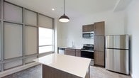 This single-floor space showcases a thoughtfully designed open-concept layout with a contemporary aesthetic. The kitchen serves as the heart of the unit, featuring rich dark wood cabinetry that extends to the ceiling for ample storage, paired with clean white countertops that provide a striking visual contrast. A central island with a smooth white surface offers additional workspace and a natural gathering point. Stainless steel appliances — including a range, built-in microwave, dishwasher, and refrigerator — lend a polished, modern finish. A classic white subway tile backsplash adds texture and detail behind the range. A standout architectural feature is the floor-to-ceiling grid-paneled window arrangement, which allows generous natural light to pour into the space, creating an airy and open atmosphere. The high ceilings further enhance the sense of volume, while a sleek pendant light fixture anchors the kitchen area with understated style. The overall ambiance is clean, modern, and functional, making excellent use of the available space through smart design choices and a cohesive neutral color palette throughout.
