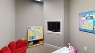 This single-floor space is thoughtfully designed with distinct functional areas connected through a series of elegant white-framed French doors with divided glass panes, creating a sense of openness while maintaining defined zones. The entryway corridor features neutral taupe walls, light beige ceramic tile flooring set in a diamond pattern, and framed botanical artwork, establishing a welcoming and polished aesthetic throughout.

The French doors open into a carpeted children's play area that serves as the centerpiece of this space. The room is outfitted with a wall-mounted flat-screen television set within a framed built-in niche, providing a clean, integrated look. Colorful educational artwork adorns the walls, including an alphabet and numbers canvas alongside a vibrant storybook-themed painting, creating a stimulating and engaging environment. A wooden book display rack holds an assortment of children's books, and a low white activity table provides a dedicated space for play and learning. The neutral gray-taupe wall tones throughout provide a calm backdrop that allows the colorful décor and furnishings to stand out, contributing to a cohesive and child-friendly atmosphere.