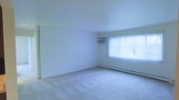 This single-floor space features a spacious, open living area defined by clean white walls and soft, light-colored carpeted flooring throughout. The room receives generous natural light through a large window fitted with vertical blinds, creating a bright and airy atmosphere. A wall-mounted air conditioning unit is installed directly above the window, providing convenient climate control. Along the base of the exterior wall, a baseboard heating system offers an additional source of warmth. The minimalist design of the space, with its smooth white ceilings and unobstructed walls, lends itself to flexible furniture arrangements and a variety of interior styles. A closet or door is visible along the left wall, adding functional storage. The overall ambiance is clean, neutral, and well-lit, offering a straightforward, practical living environment.